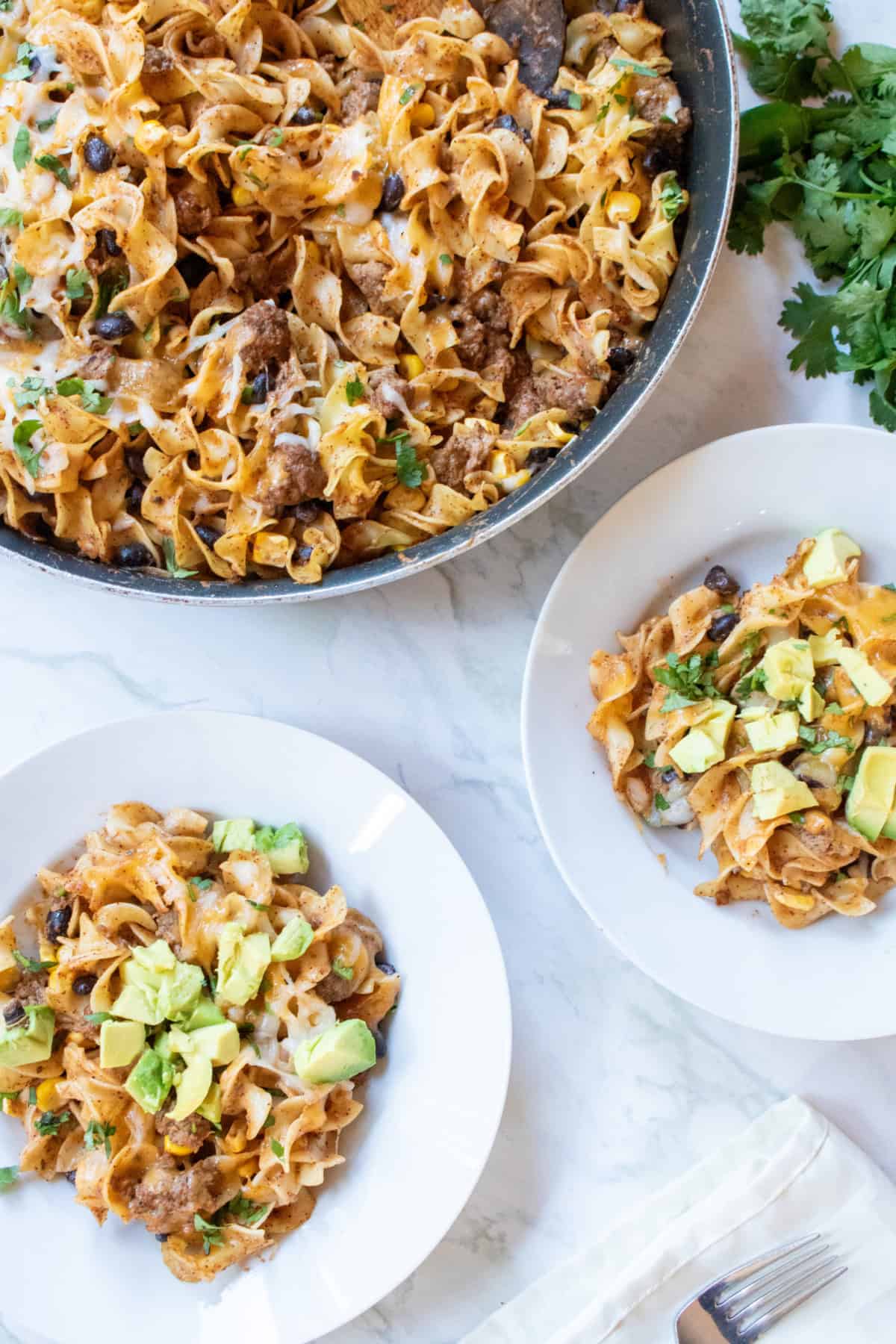 an overhead of a cheesy pasta skillet with beef and beans and two bowls of the pasta topped with diced avocado.