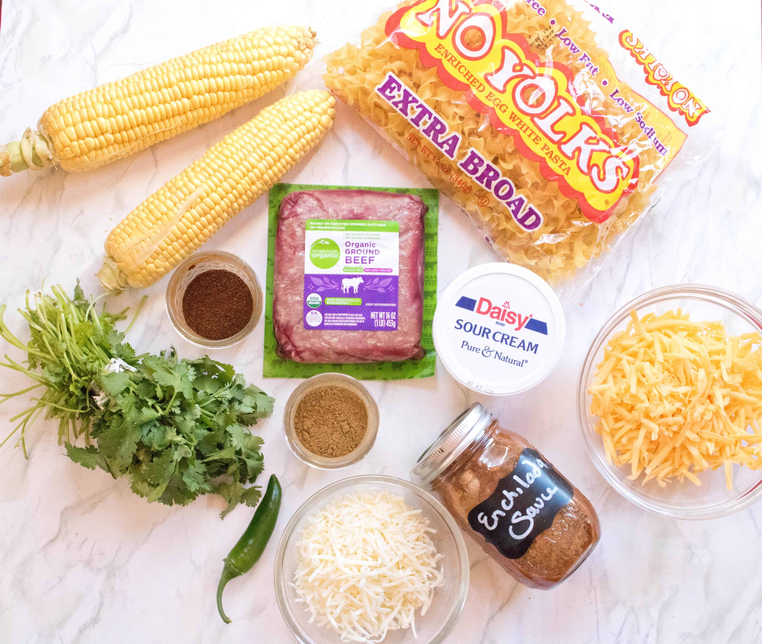 noodles, beef, corn, cheese, enchilada sauce, spices, sour cream, and cilantro on counter.