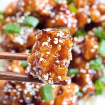 cooked crispy sesame chicken held up by chopsticks.