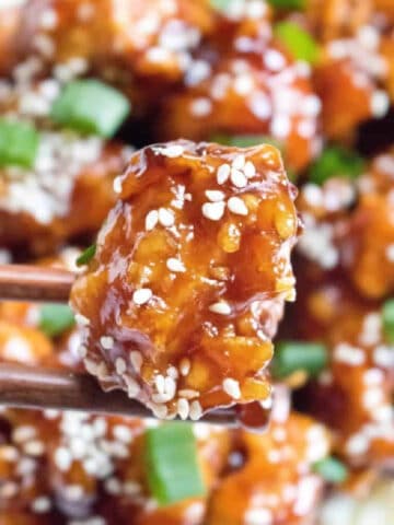 cooked crispy sesame chicken held up by chopsticks.