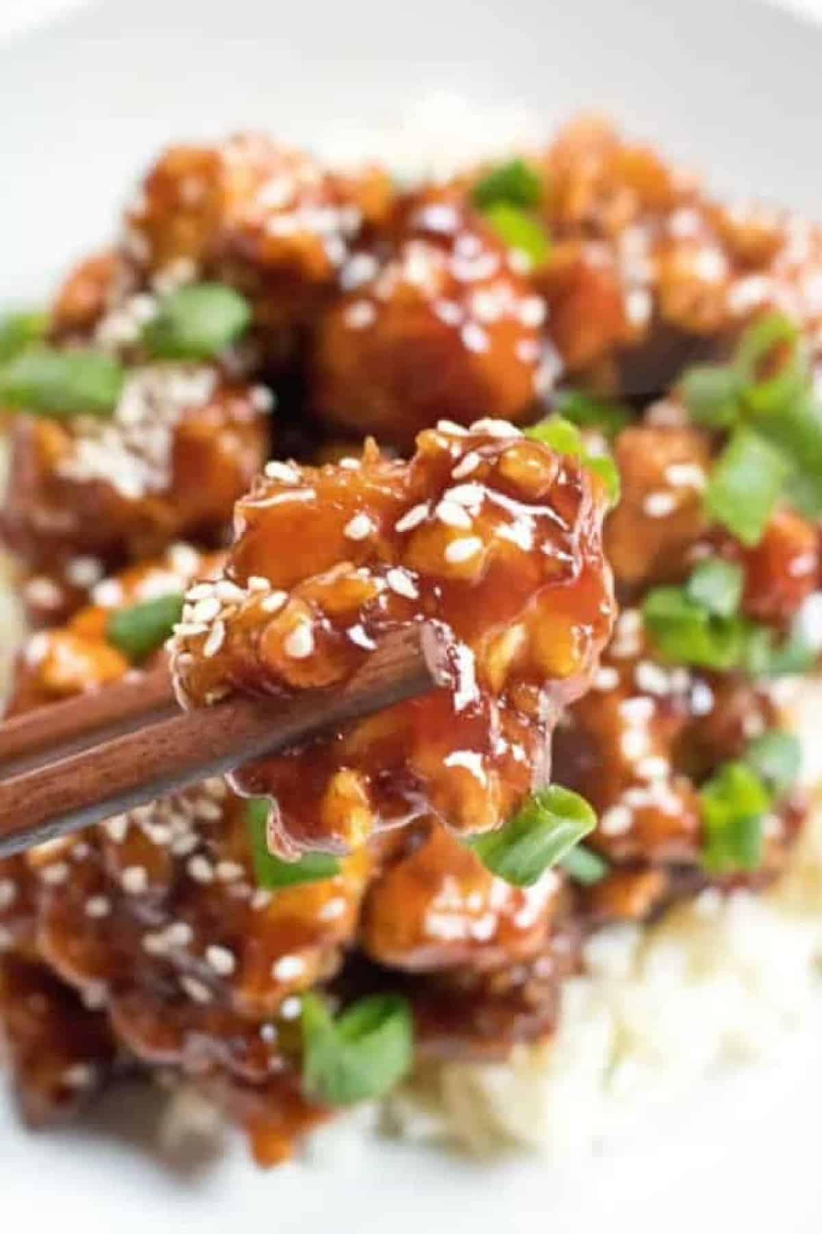 cooked crispy sesame chicken held up by chopsticks.