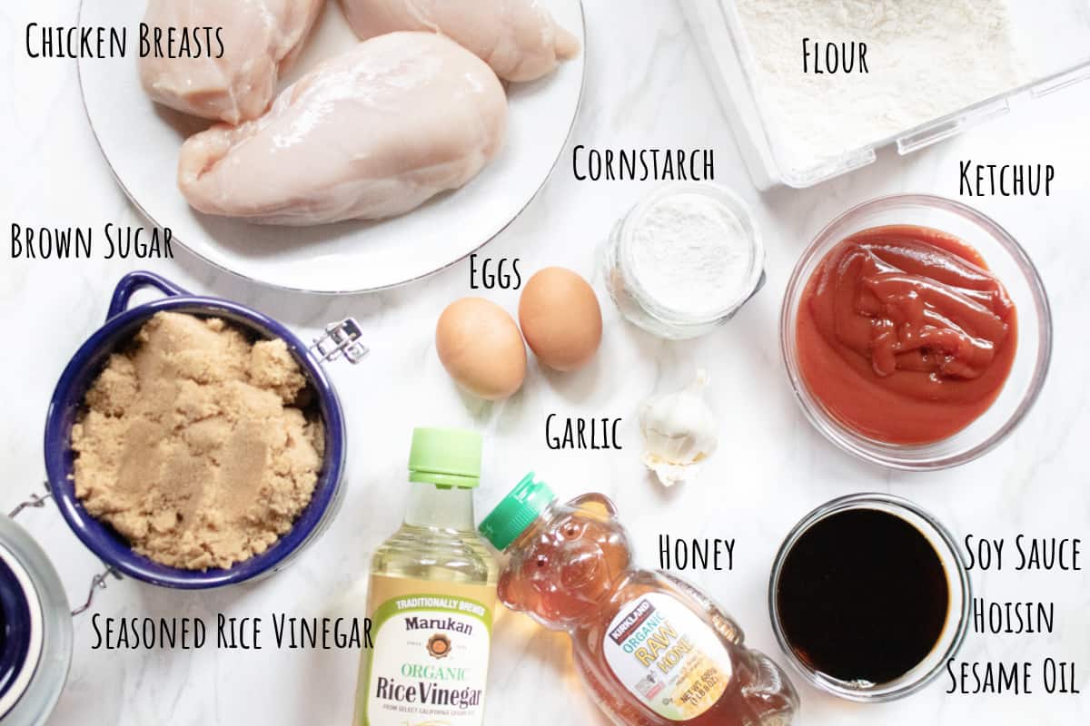 raw chicken breasts, flour, ketchup, cornstarch, eggs, garlic, soy sauce, honey, rice vinegar, brown sugar on a counter.