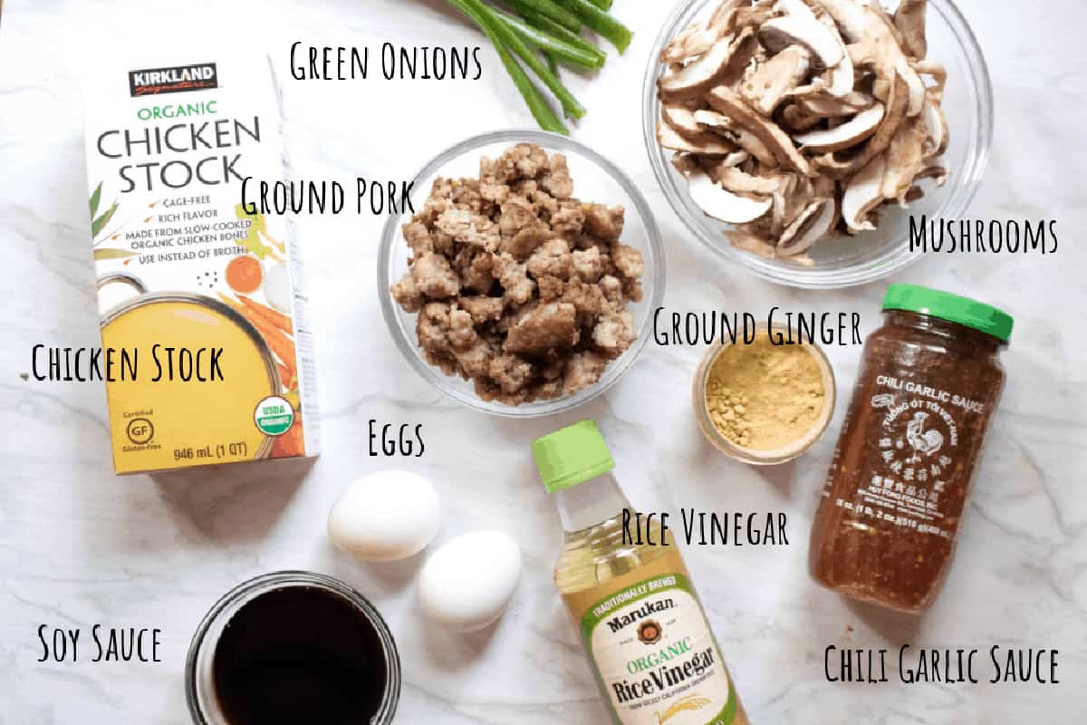 chicken stock, soy sauce, ground cooked pork, mushroom, garlic chili sauce, rice vinegar, eggs, green onions, and ginger on a counter.