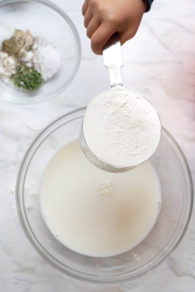 a toddler hand holding a cup of flour over a bowl of milk with spices next to it.