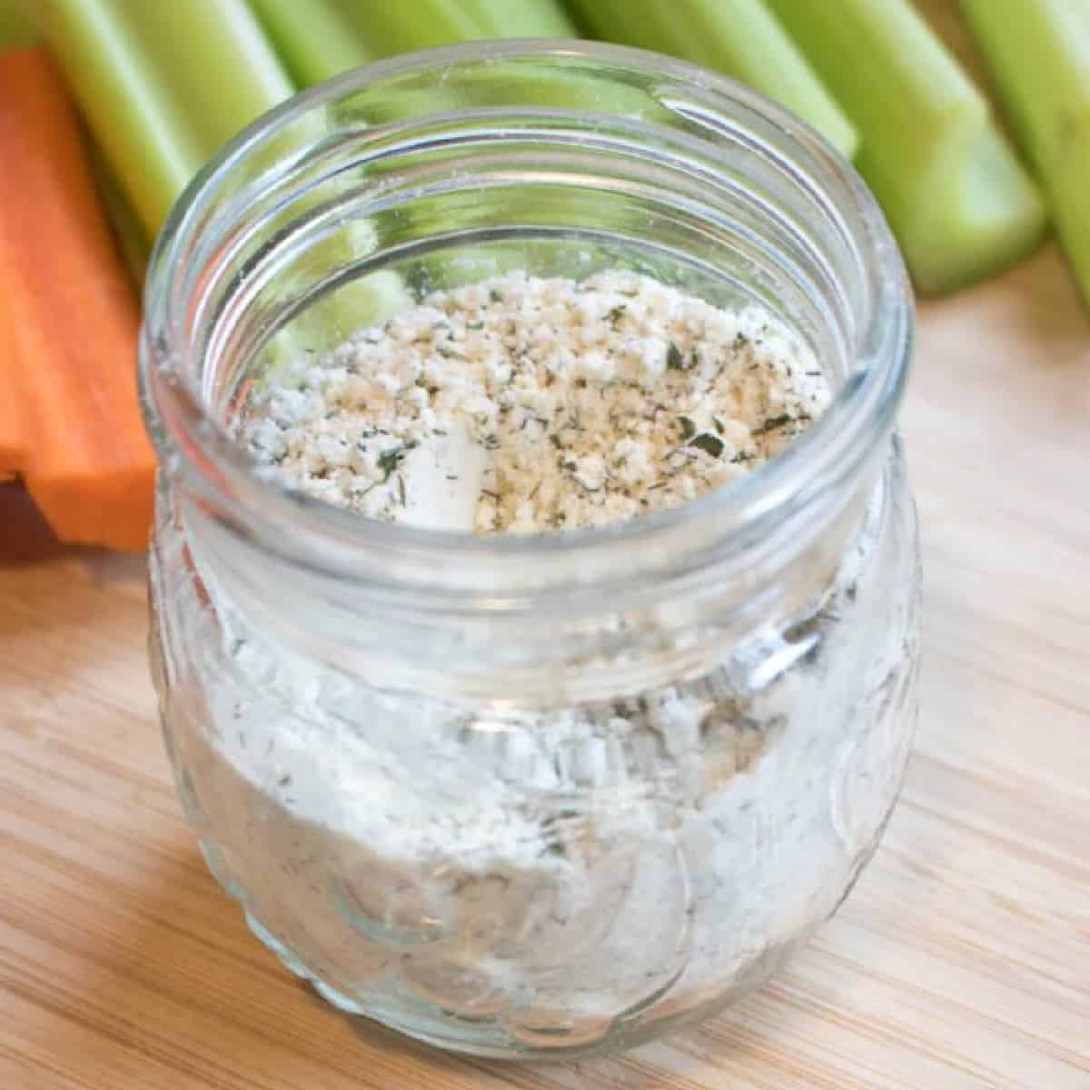 a small jar with dry ranch mix and a measuring spoon on a cutting board with carrots and celery