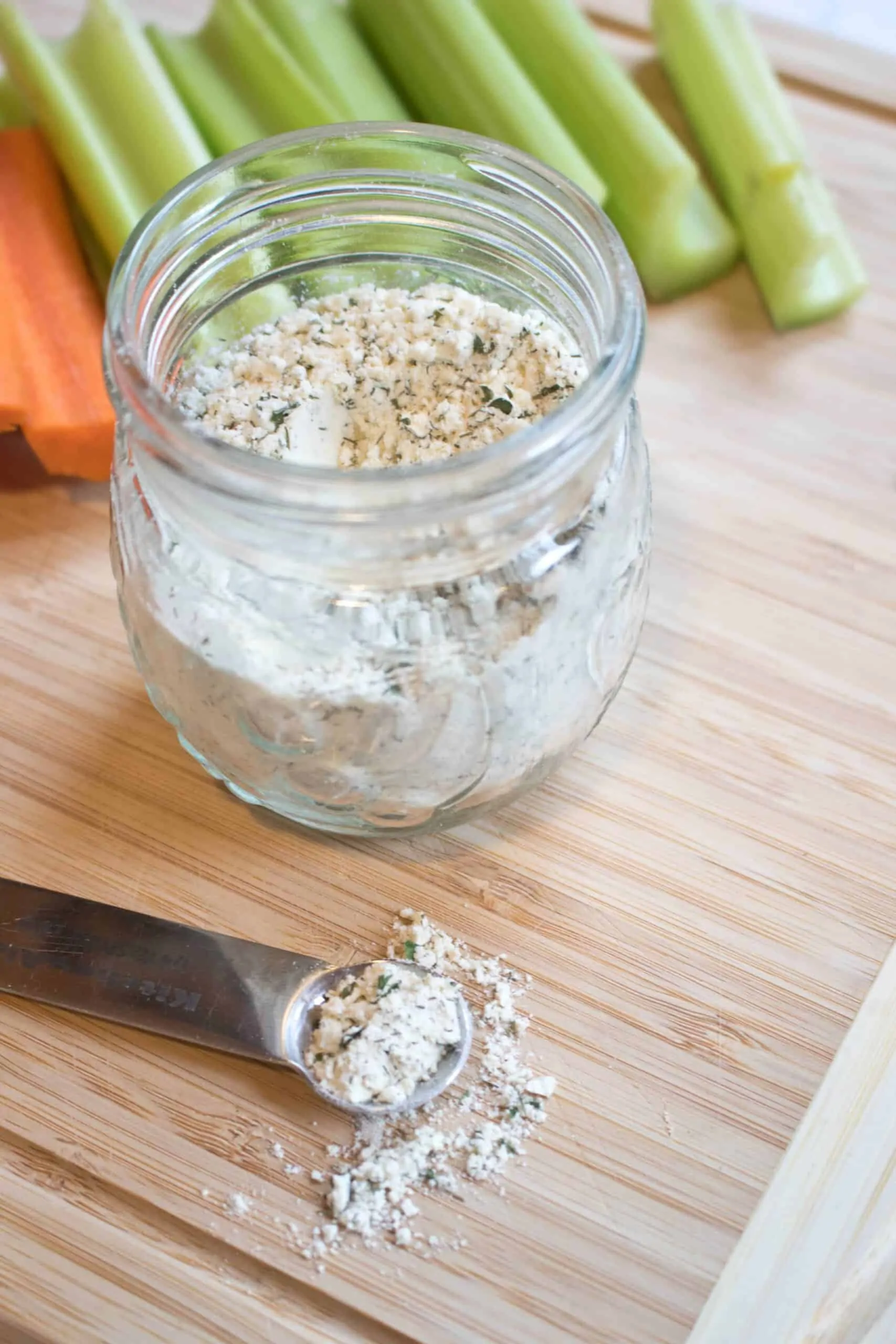 a small jar with dry ranch mix and a measuring spoon on a cutting board with carrots and celery