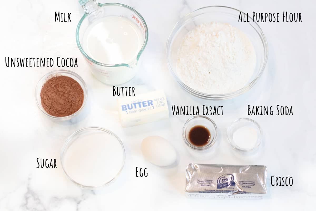 milk, flour, sugar, cocoa, baking soda, vanilla extract, crisco, egg and butter on a counter.