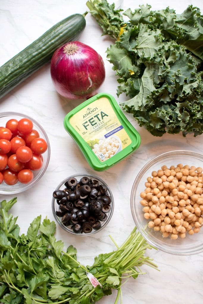 Tomatoes, Feta, olives, cucumbers, tomatoes, kale, and chickpeas and onion on a counter.