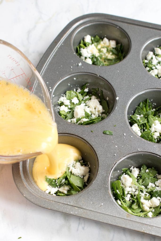 muffin tin with spinach and feta with eggs pouring it in.