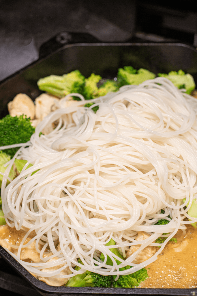 rice noodles in a pan with broccoli and chicken.