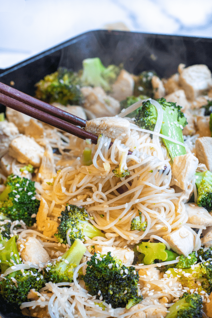 a cast iron with noodles, broccoli, and chicken with sesame seeds and chopsticks.