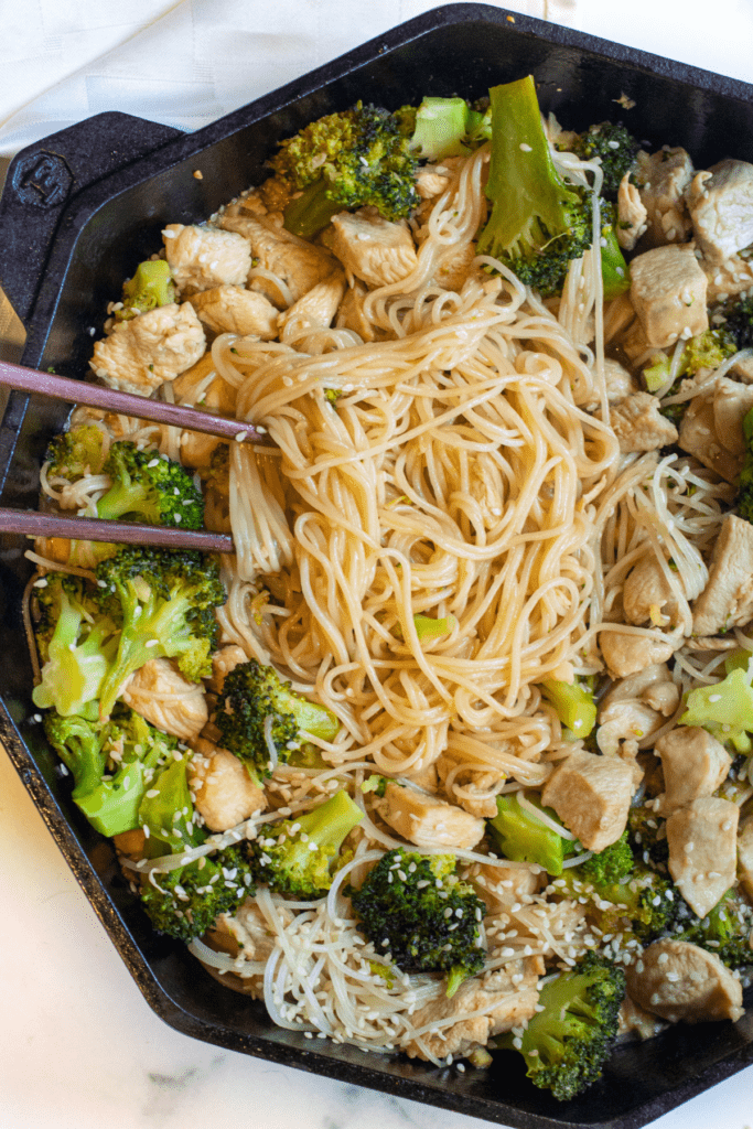 a cast iron with noodles, broccoli, and chicken with sesame seeds and chopsticks.