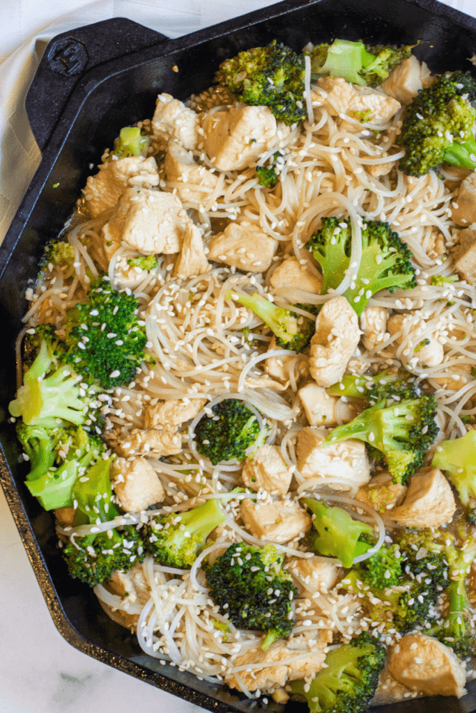 a cast iron with noodles, broccoli, and chicken with sesame seeds.