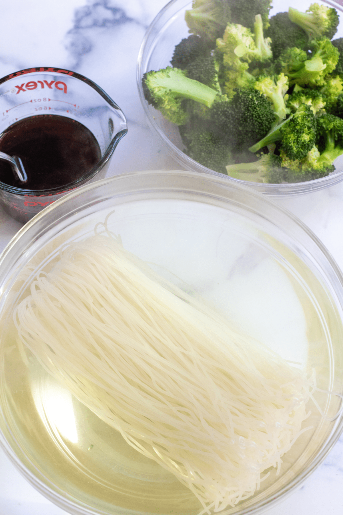 a bowl of rice noodles, cooked broccoli florets, and sauce.