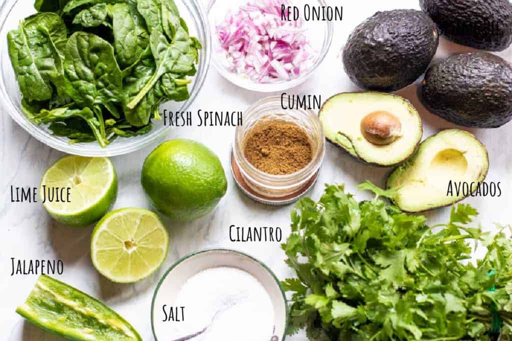 spinach, avocados, onions, cilantro, limes, cumin, salt, jalapeno on a counter.