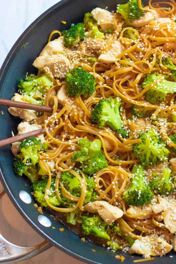 a pan of cooked noodles, broccoli, chicken topped with sesame seeds