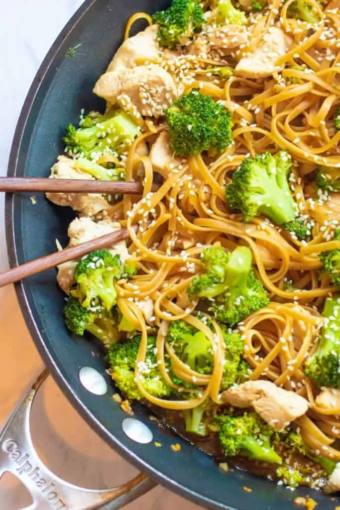 a pan with noodles, broccoli, chicken, topped with sesame seeds