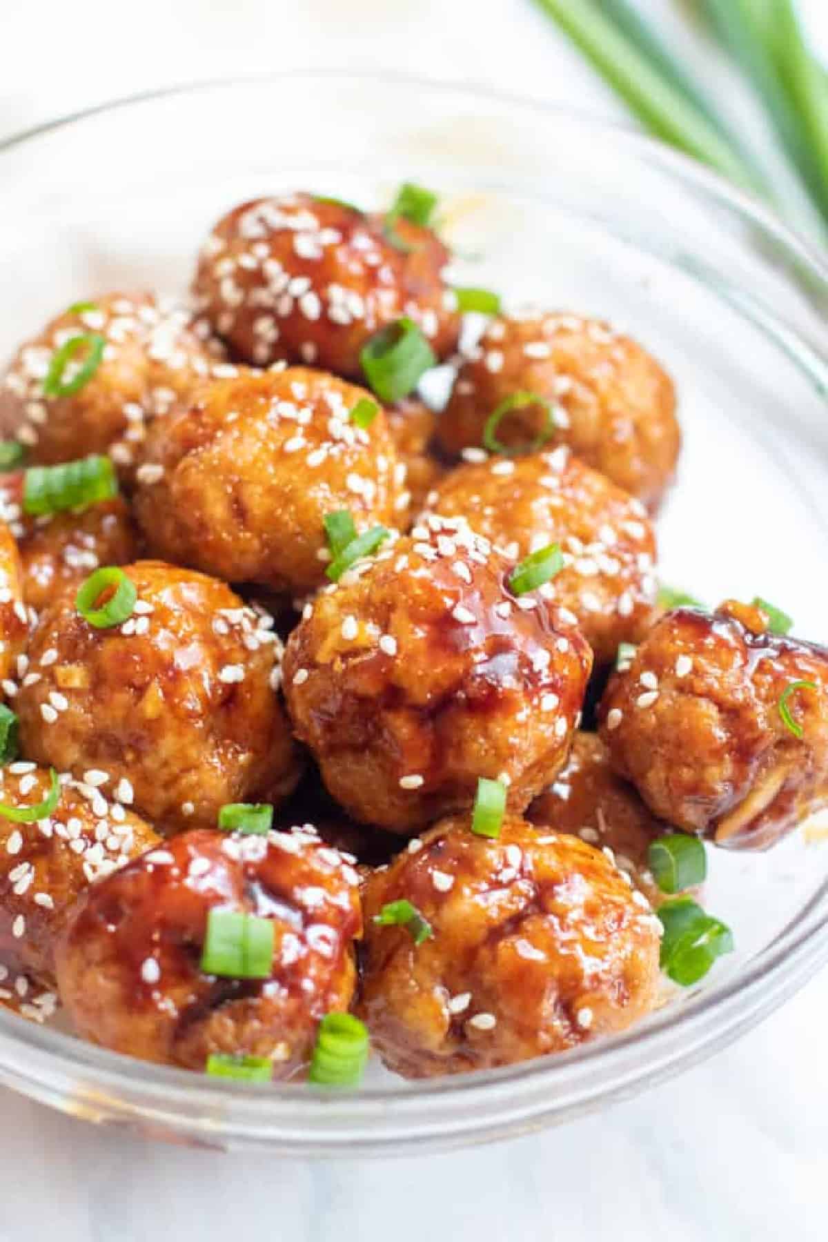 a bowl of sauced meatballs with sesame seeds and green onions.