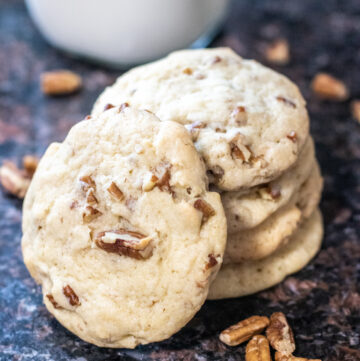 stacked pecan cookies on a counter with milk and pecan pieces.