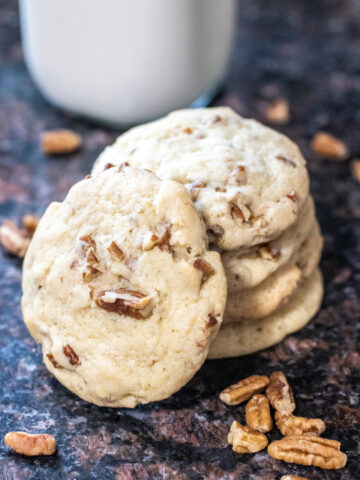 stacked pecan cookies on a counter with milk and pecan pieces.