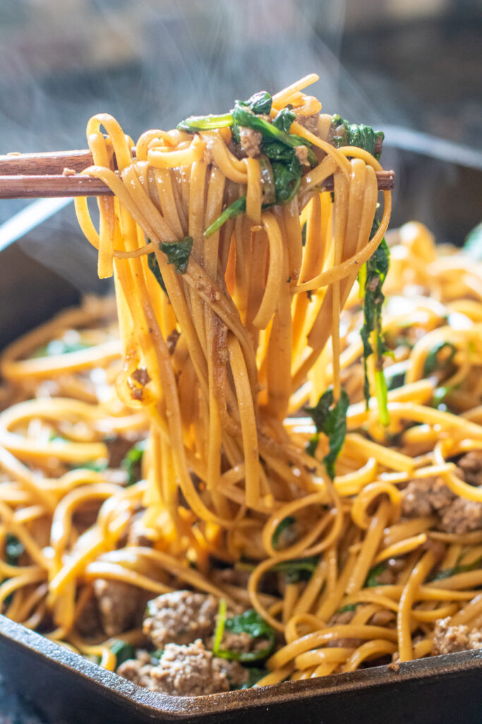 chopsticks holding cooked noodles with spinach and ground beef from cast iron