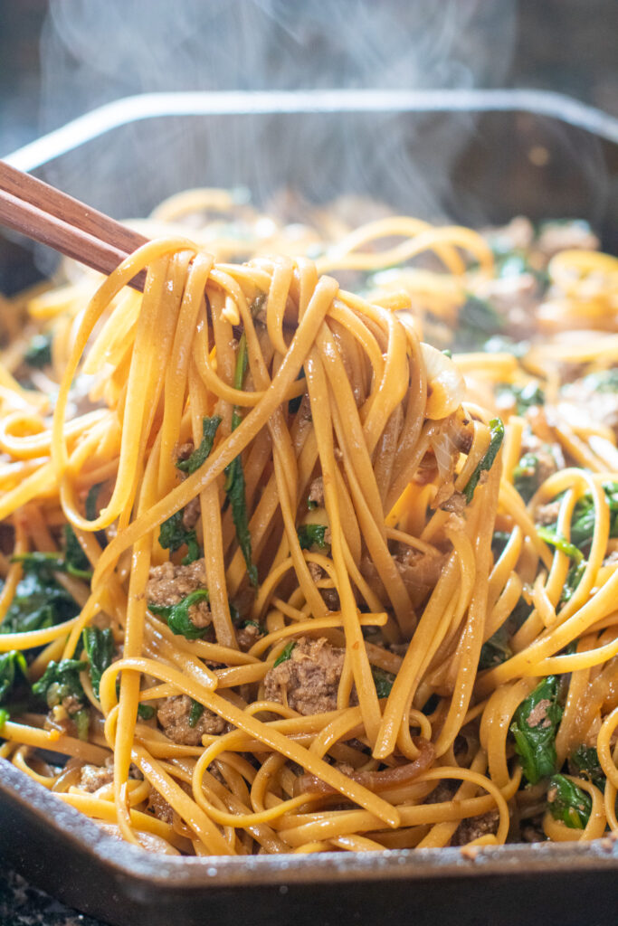 chopsticks holding up cooked noodles with beef and spinach in cast iron.