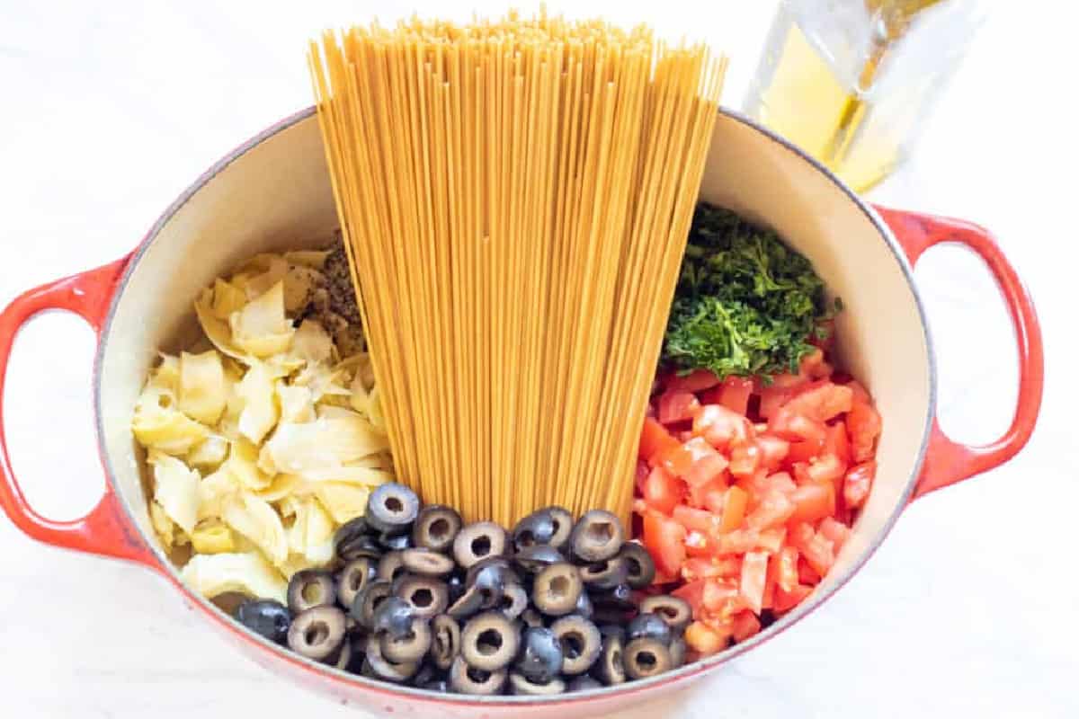 a dutch oven with dry pasta, olives, tomatoes, fresh herbs, and artichokes.