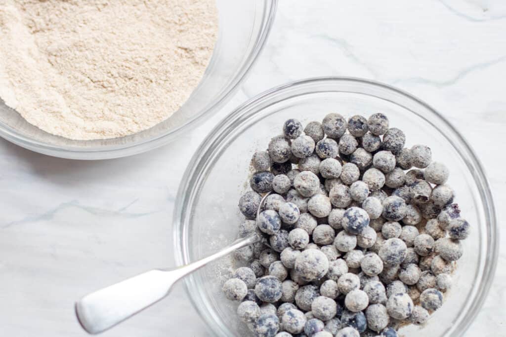 a bowl of flour and a bowl of flour coated blueberries with a spoon