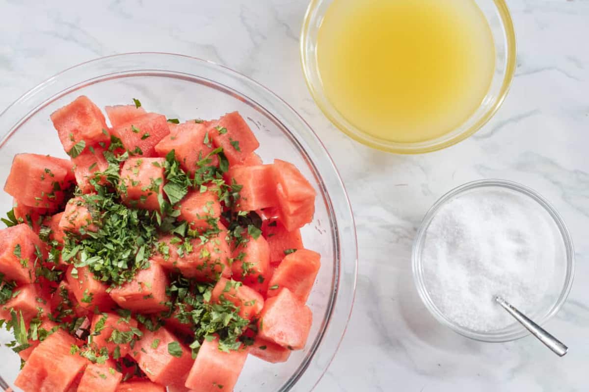 a bowl of cubed watermelon with mint and a bowl of juice and a bowl of salt.