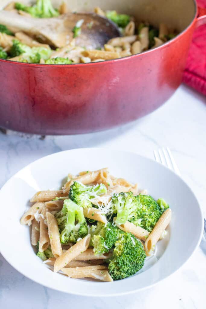 a plate of cooked penne and broccoli in front of a red pot