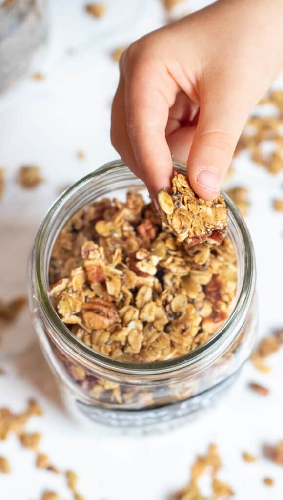 a kid's hand holding a cluster of granola over a mason jar 