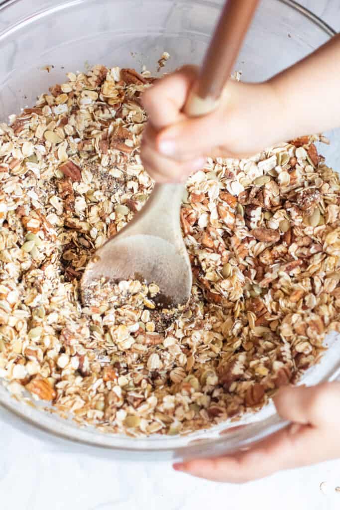 A large bowl with oats, nuts, seeds, being mixed with a wooden spoon and toddler hand