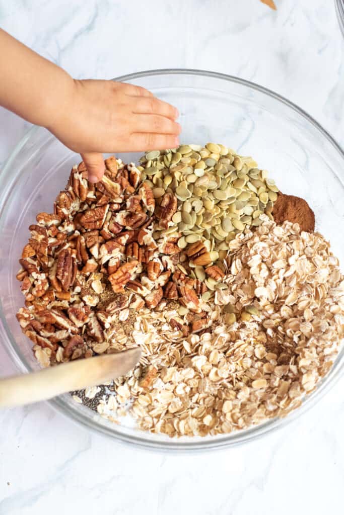 a large bowl with oats, nuts, seeds, and cinnamon with a wooden spoon and a toddler hand