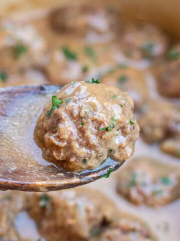 a wooden spoon holding up a swedish meatball over a pot of meatballs.