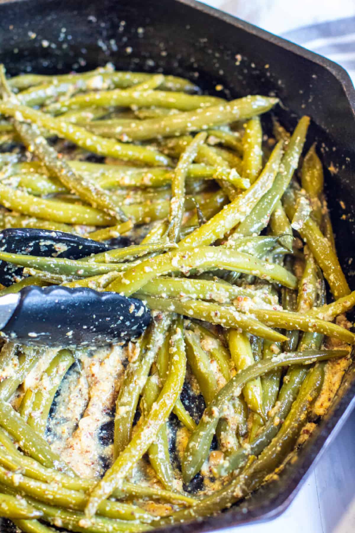 cooked green beans in a pan coated with mustard with tongs.