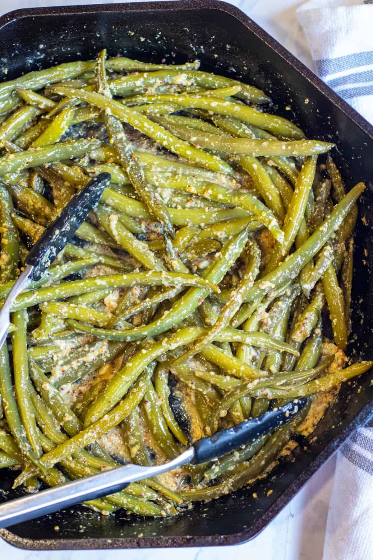 cooked green beans in a pan coated with mustard with tongs.