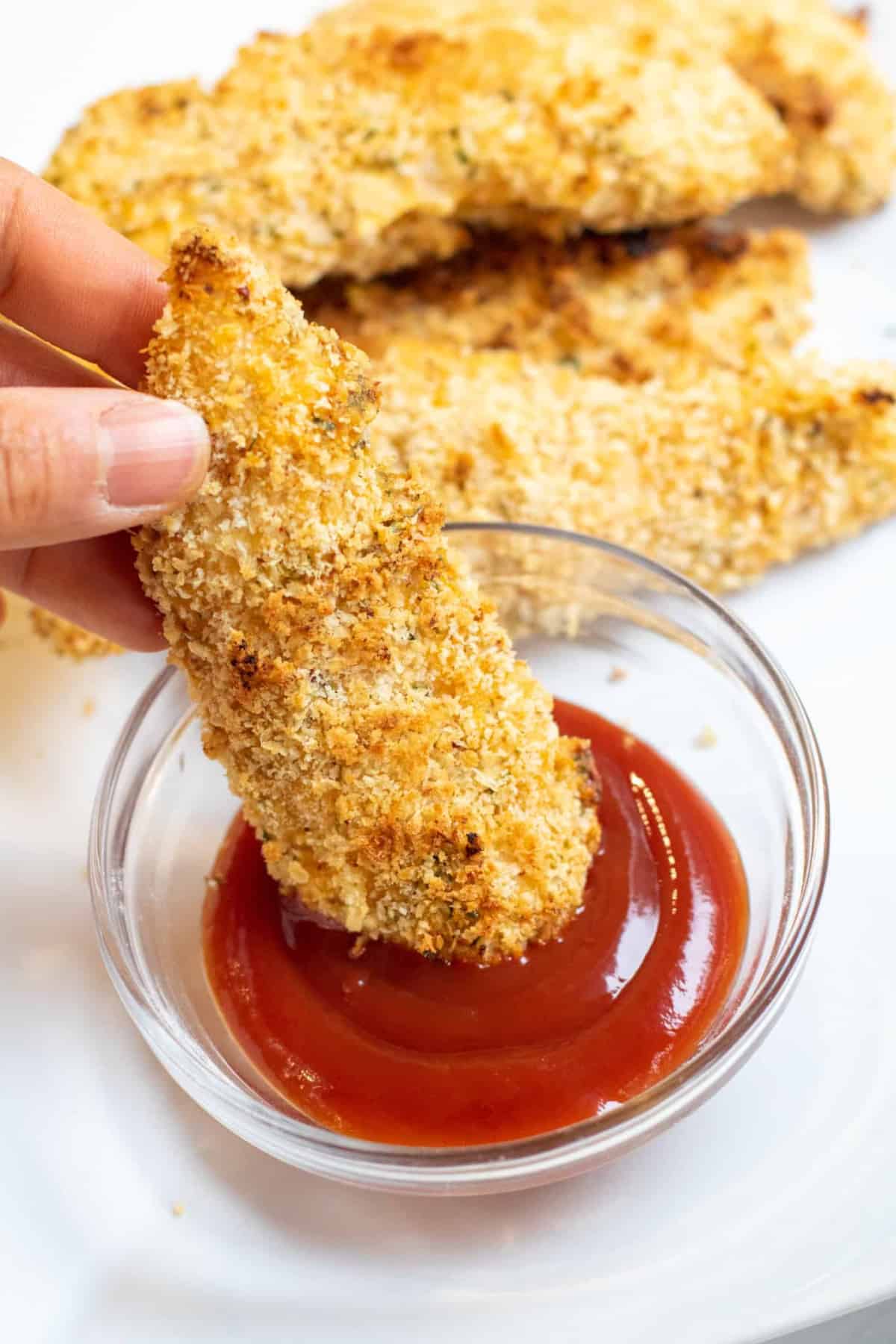 a hand dipping a crispy chicken tender into ketchup.