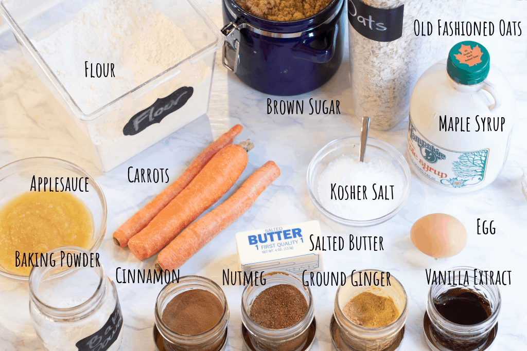 flour, applesauce, sugar, oats, carrots, maple syrup, salt, egg, baking soda, cinnamon, nutmeg, ginger, and vanilla extract on a counter