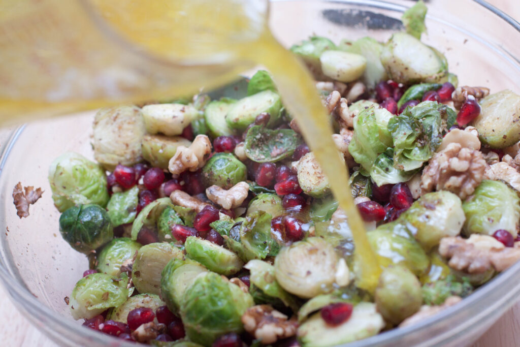 a bowl of brussels sprouts salad with pomegranates and cheese with vinaigrette being poured in.