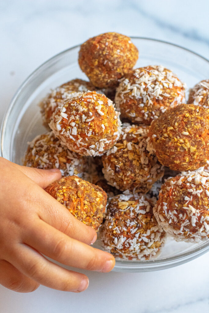 a bowl of oat and carrot balls with coconut with toddler hand grabbing one.