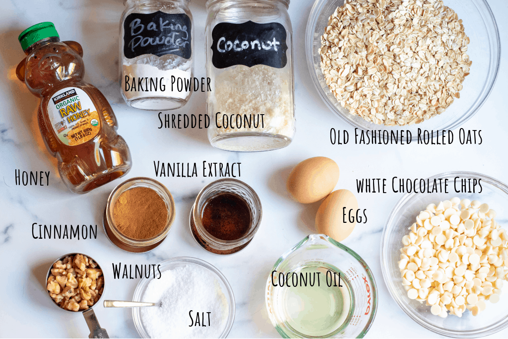 oats, coconut, baking powder, honey, cinnamon, vanilla, eggs, melted coconut oil, walnuts, salt, white chocolate chips on counter.