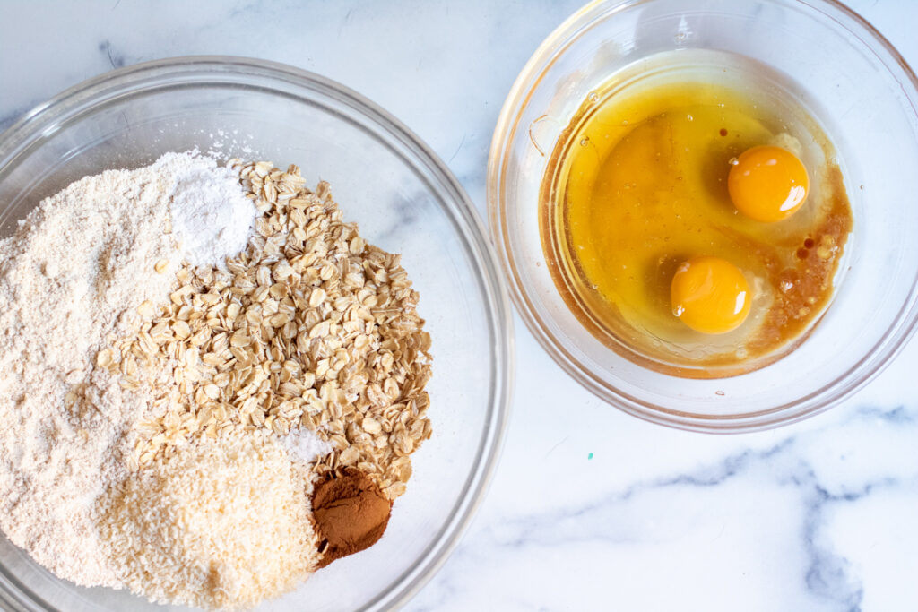 a bowl with oats, oat flour, baking powder, cinnamon, salt, next to a bowl of eggs, vanilla, and coconut oil.