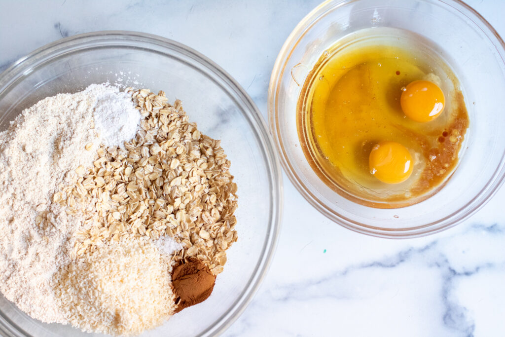 a bowl with oats, oat flour, baking powder, cinnamon, salt, next to a bowl of eggs, vanilla, and coconut oil.