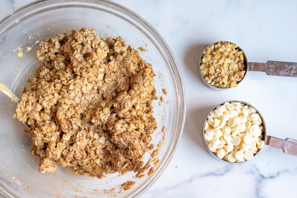 a bowl of oatmeal cookie dough with measuring cups of chocolate chips and walnuts.