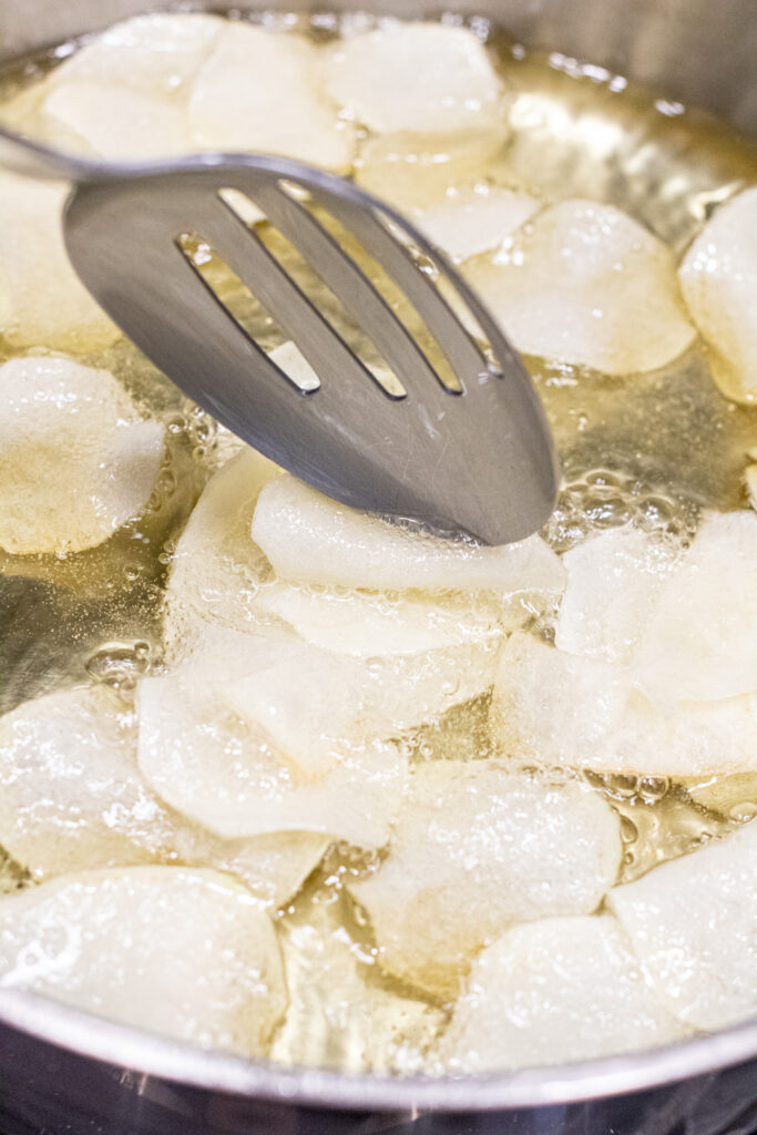 a slotted spoon in a pan of oil and sliced potatoes folding a potato.