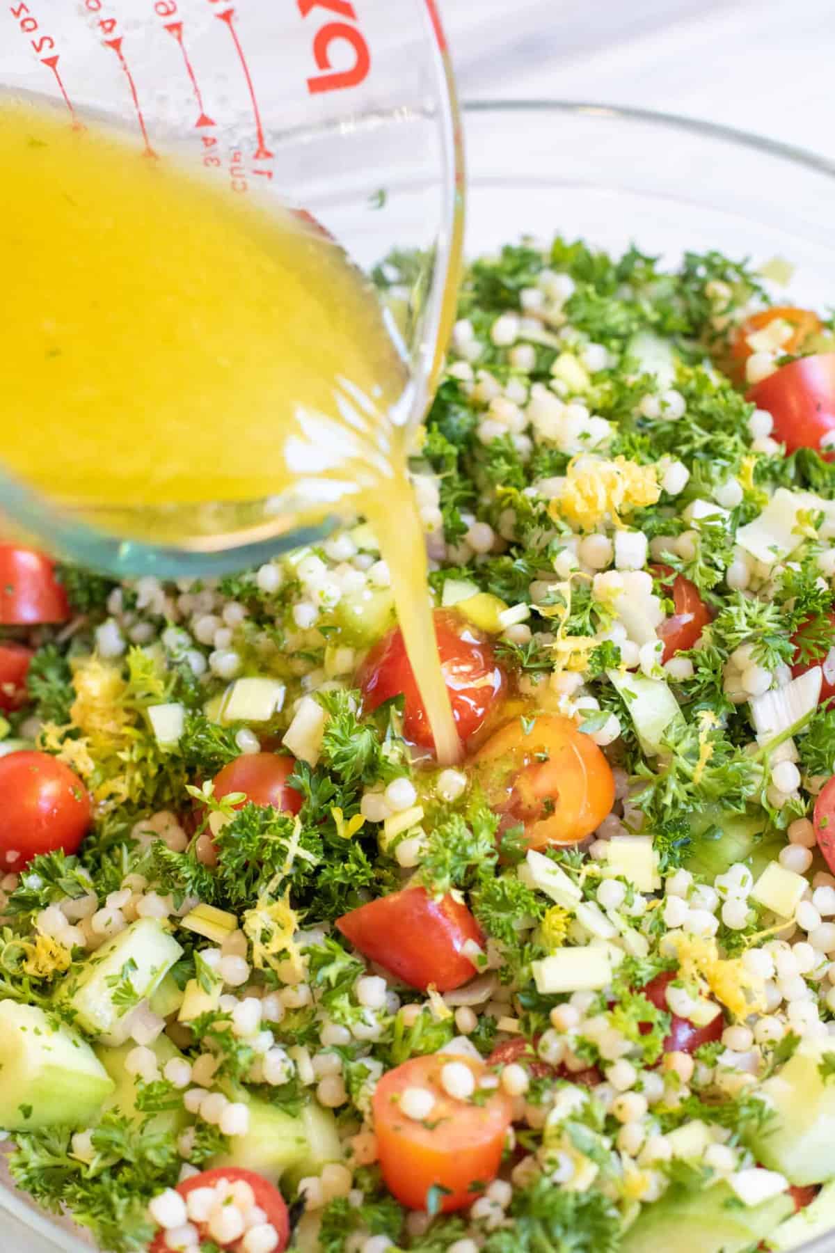 a bowl of couscous, cucumbers, tomatoes, parsley with vinaigrette being poured in.