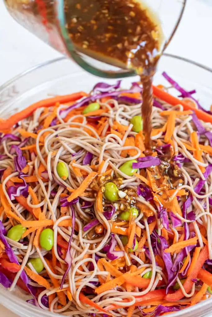 dressing being poured into bowl of sesame noodle with carrots, cabbage, and edamame.