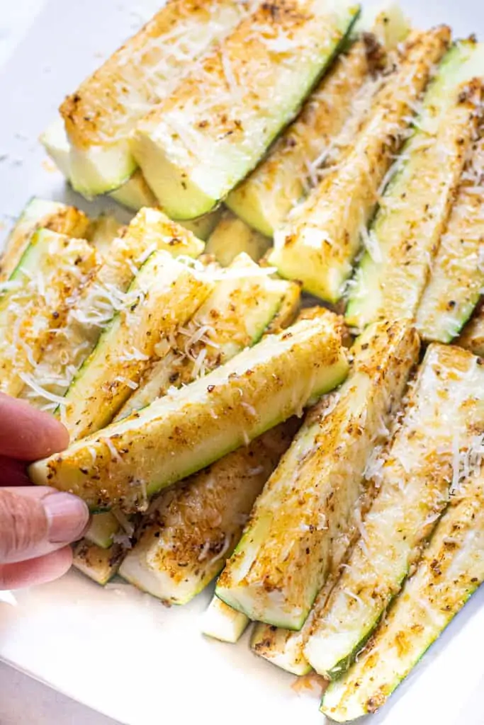 a hand grabbing a zucchini spear from a plate of baked zucchini spears.
