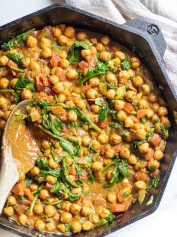 a cast iron of chickpea curry with spinach with a wooden spoon.