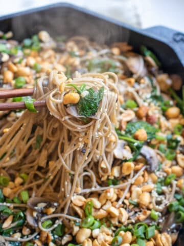 cast iron of noodles with spinach, mushrooms, topped with peanuts and scallions chopsticks holding up noodles.